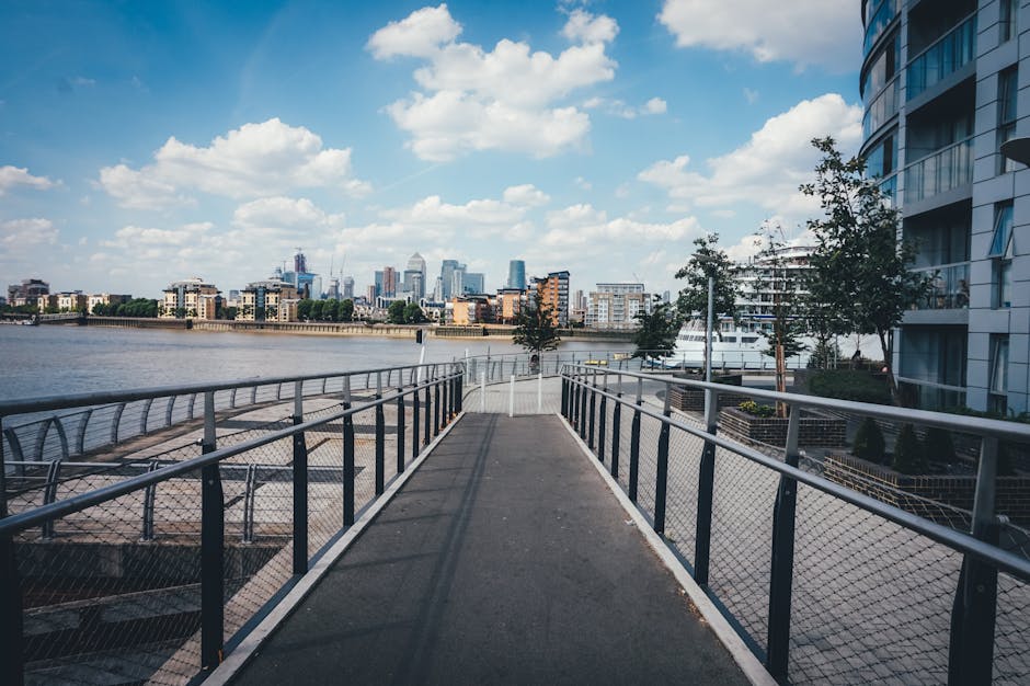 Awide, straight pathway constructed of dark, textured material, bordered by metal railings with mesh panels, extends towards a river with calm water reflecting the partly cloudy sky. On one side of th