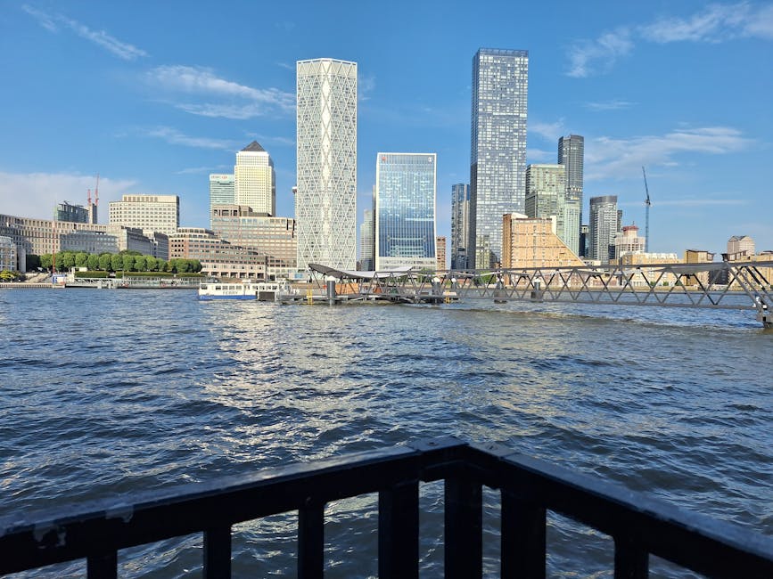 Awide, straight pathway constructed of dark, textured material, bordered by metal railings with mesh panels, extends towards a river with calm water reflecting the partly cloudy sky. On one side of th