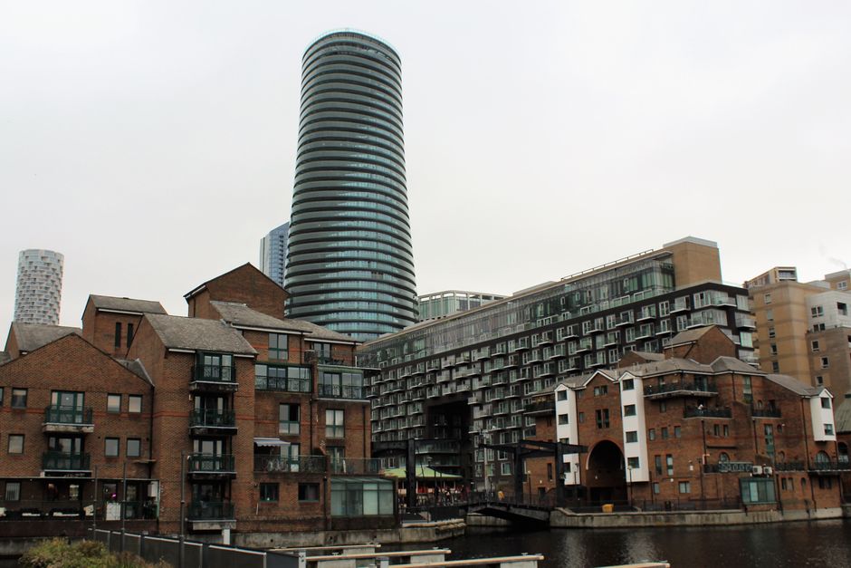 A cityscape featuring a cluster of modern and historic buildings with a focus on a group of red-brick, multi-storey townhouses with small balconies and pitched roofs. Behind these, there are contempor