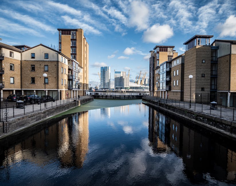 A cityscape featuring a cluster of modern and historic buildings with a focus on a group of red-brick, multi-storey townhouses with small balconies and pitched roofs. Behind these, there are contempor